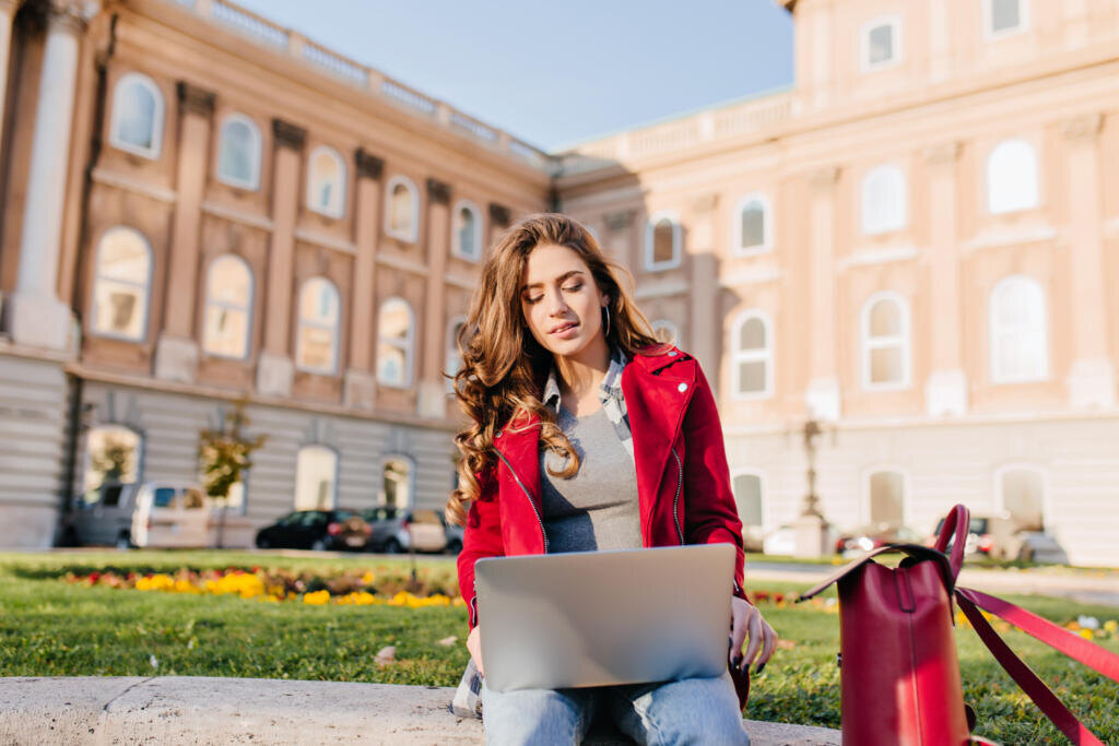 Outdoor portrait of serious curly female student sitting with laptop on the ground. Busy brunette girl in red jacket working with computer in front of college in warm day. Журналист