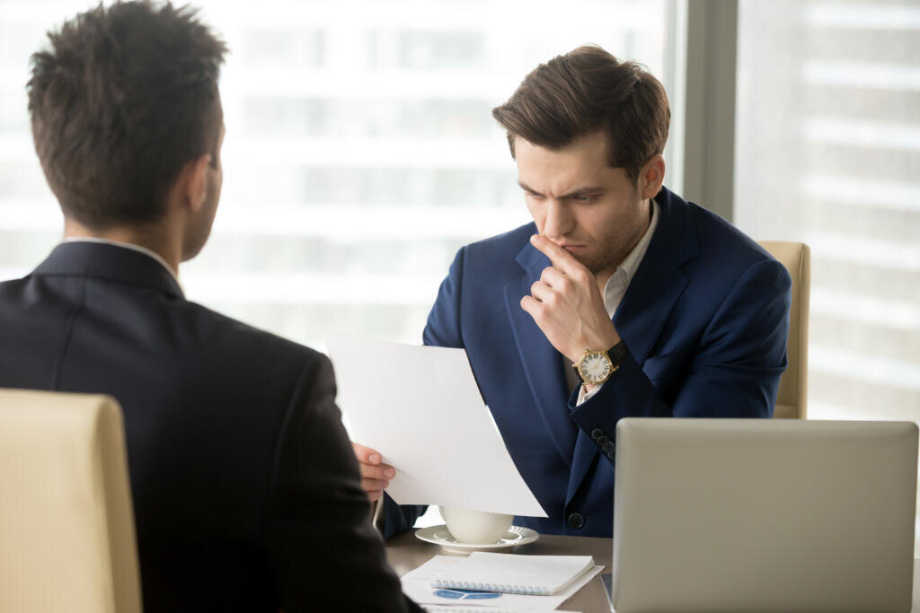 Doubting businessman reading contract document with suspicion when sitting at desk in front of business partner. Entrepreneur unsure in terms of agreement, feeling skeptical because of financial plan Журналист