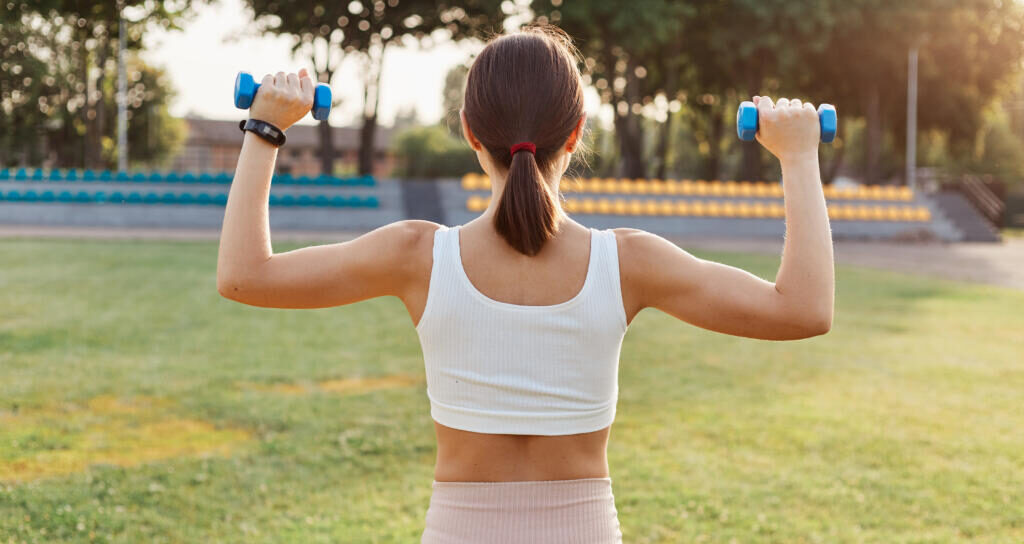 Back view of dark haired female with sporty body holding dumbbells and doing exercises on stadium, training biceps and triceps, outdoor activity, healthy lifestyle. Журналист