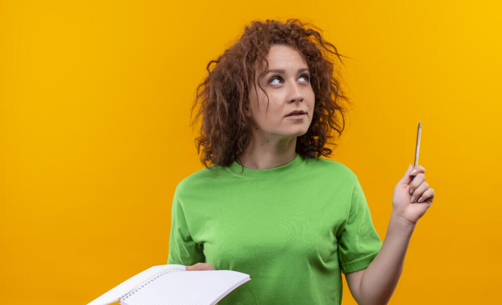 young woman with short curly hair in green t-shirt holding notebook and pen looking up thinking standing over orange background Журналист