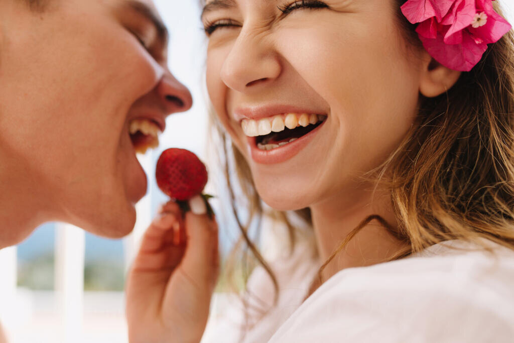 Excited happy young woman with cute pink flower in light-brown hair feeding her laughing husband with fresh strawberry. Close-up portrait of romantic enjoying honeymoon and eating berries Журналист