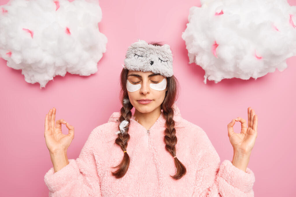 Relaxed beautiful woman in pajamas meditates after awakening keeps hands in zen gesture closes eyes enjoys peaceful atmosphere undergoes beauty treatments at home practices yoga stands indoor Журналист