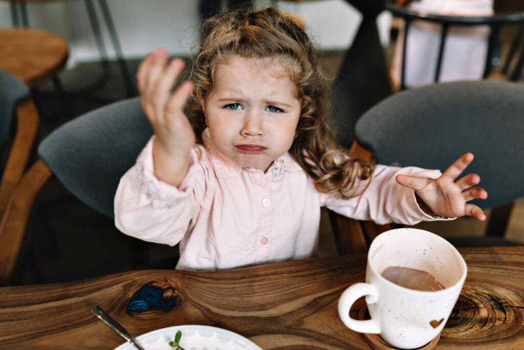 Funny little girl is sitting at a table in a restaurant, holding up hand and looking at camera. She is sitting with cacao and dessert and playing while dinner Журналист