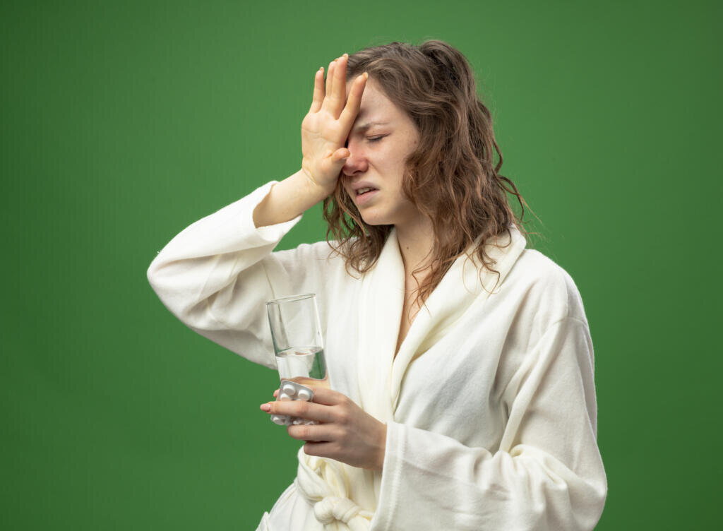 weak young ill girl with closed eyes wearing white robe holding glass of water putting hand on forehead isolated on green background Журналист