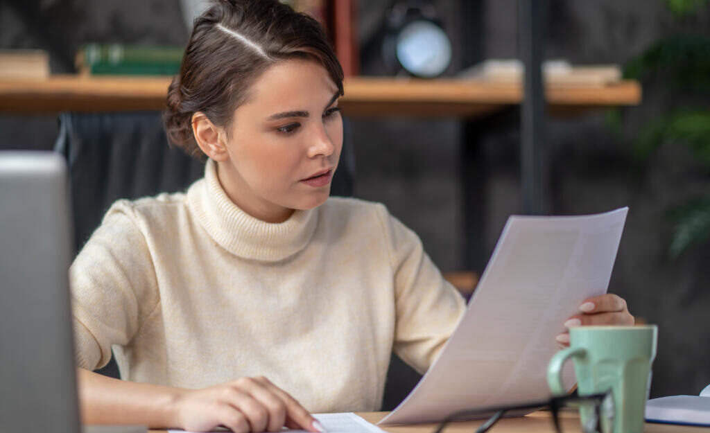 Focused young Caucasian woman sitting at the writing table while scrutinizing documents in her hand Журналист
