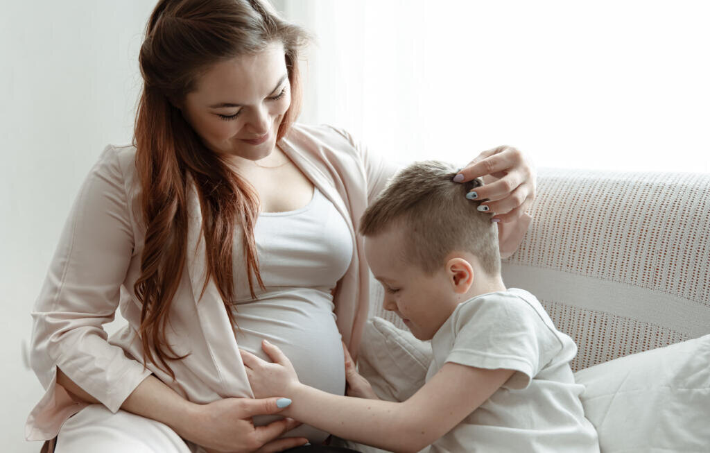 Child boy hugging the belly of pregnant her mother on the couch at home. Журналист