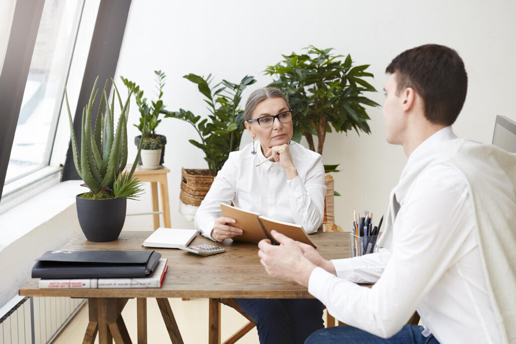 People, work, employment, occupation and profession concept. Experienced serious gray haired mature female human resources specialist holding notebook while conducting job interview with young man Журналист