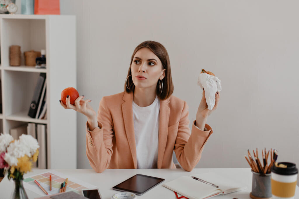 Portrait of lady with short hair in office. Business woman in thought chooses to eat burger or healthy apple Журналист