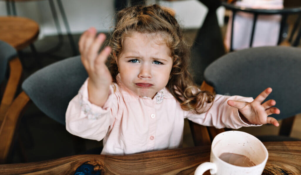 Funny little girl is sitting at a table in a restaurant, holding up hand and looking at camera. She is sitting with cacao and dessert and playing while dinner Журналист