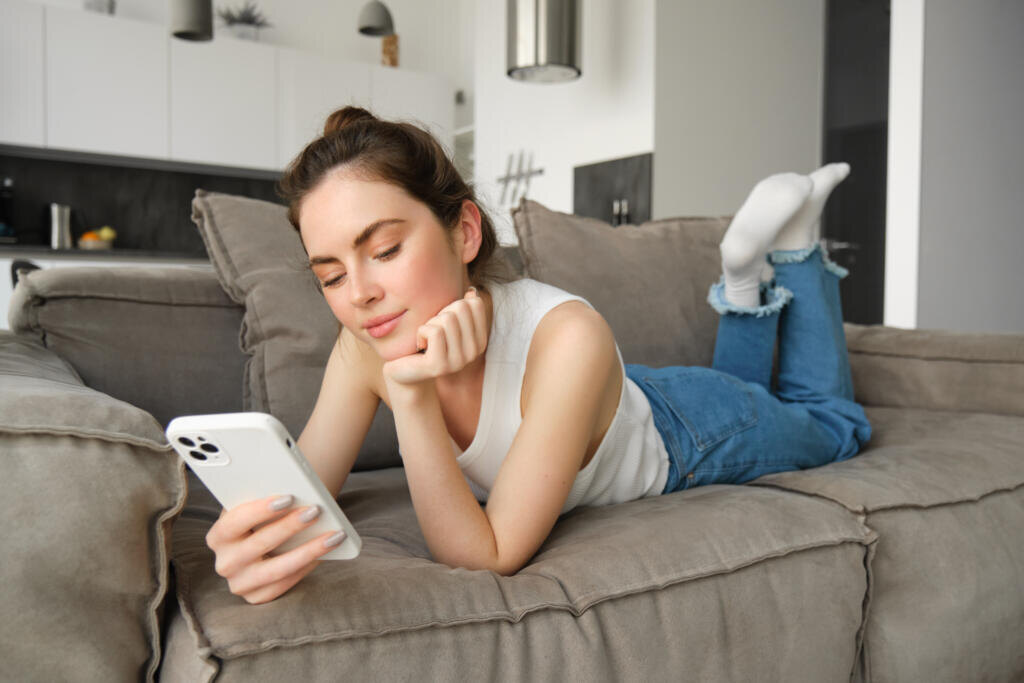 Portrait of smiling young woman lying on sofa at home, looking at her mobile phone, reading message on social media, using smartphone on couch. Журналист