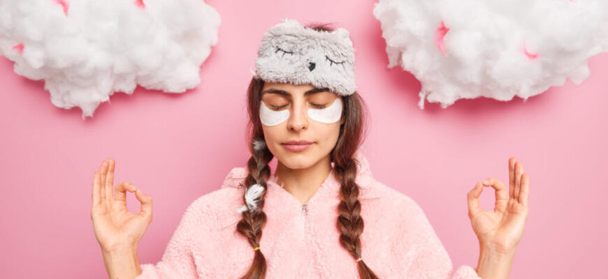 Relaxed beautiful woman in pajamas meditates after awakening keeps hands in zen gesture closes eyes enjoys peaceful atmosphere undergoes beauty treatments at home practices yoga stands indoor Журналист