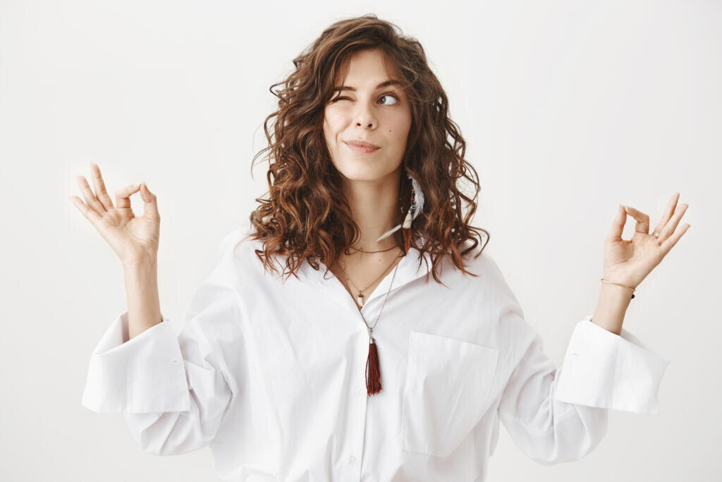 Studio portrait of charming positive caucasian female businesswoman trying to relax while meditating, standing with lifted hands and zen signs over gray background, peeking and looking aside playfully. Журналист
