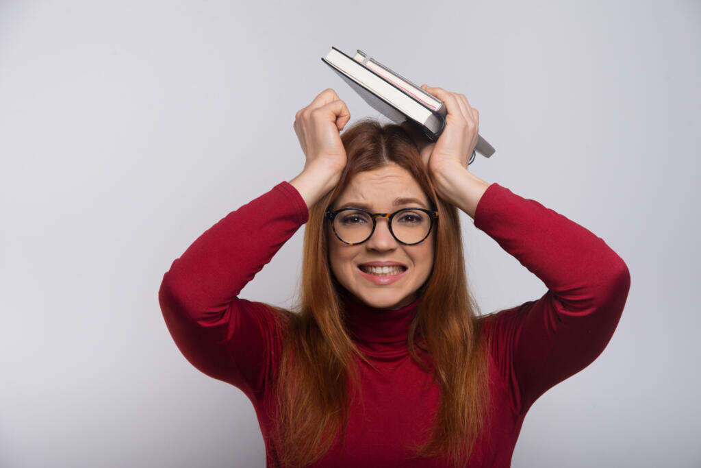 Stressed female student with books over head. Tired young woman in eyeglasses holding textbooks and looking up isolated on white background. Education concept Журналист