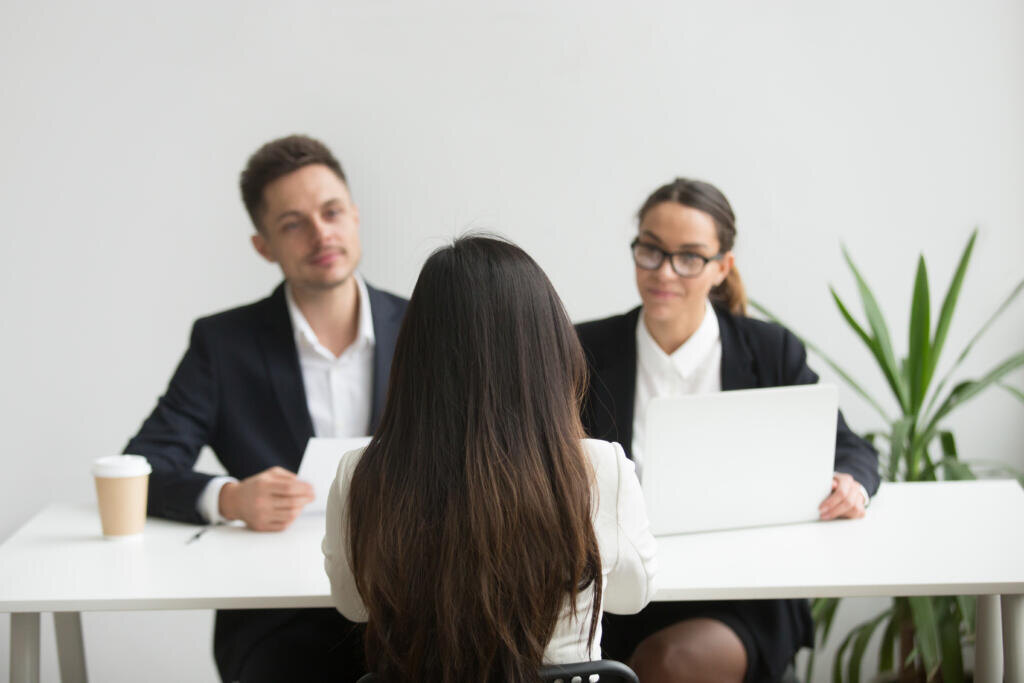 Back close up view of female applicant being interviewed by two HR managers reading her resume, checking data on laptop, asking questions for job position. Employment, hiring, first impression concept Журналист
